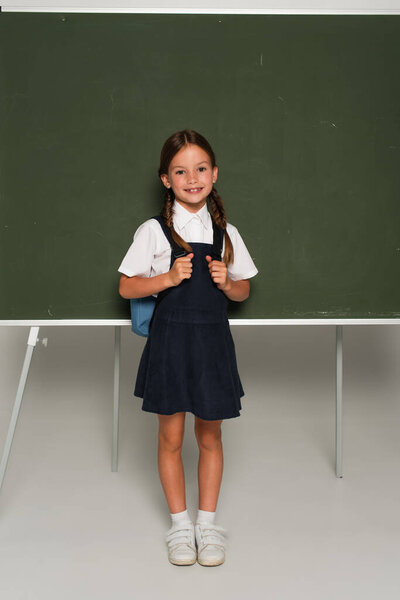 full length view of schoolchild with blue backpack smiling at camera near chalkboard on grey
