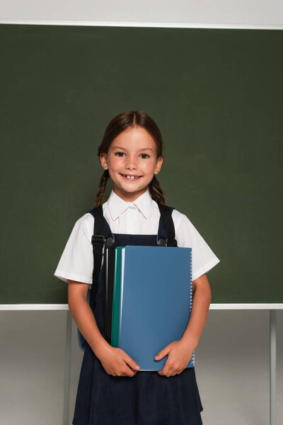 smiling schoolkid standing near chalkboard with notebooks on grey