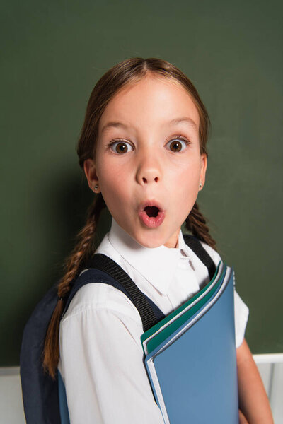thrilled schoolchild with copy books looking at camera near chalkboard on grey