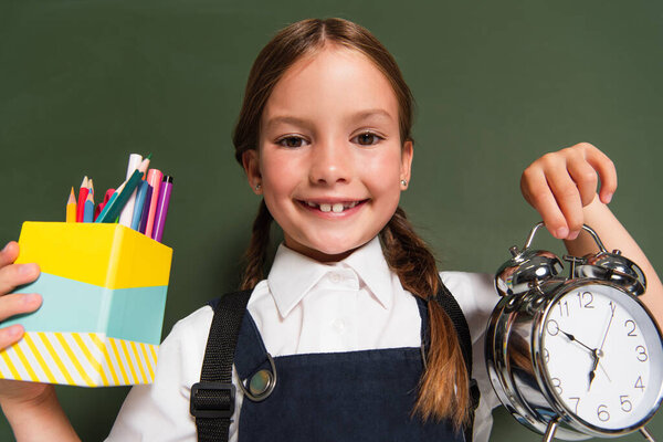 joyful schoolgirl showing pen holder and alarm clock near chalkboard
