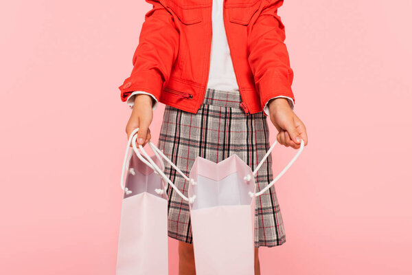 cropped view of child in orange jacket and plaid skirt opening shopping bag isolated on pink