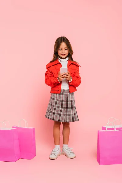 穿着橙色夹克和格子裙的孩子在粉色购物袋附近用智能手机 — 图库照片
