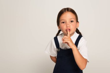 girl in school uniform showing hush sign while looking at camera isolated on grey clipart