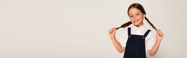 Радостная школьница смотрит в камеру, прикасаясь к косичкам, изолированным на сером, баннер