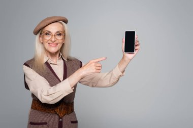 Gülümseyen yaşlı kadın gri ekranda boş ekranlı cep telefonunu işaret ediyor. 