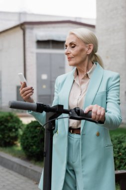 Mavi ceketli yaşlı bir iş kadını elektrikli scooter 'ın yanındaki cep telefonuna bakıyor.