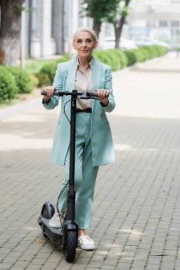 Şık takım elbiseli üst düzey bir iş kadını şehrin caddesinde elektrikli scooterıyla dikiliyor.