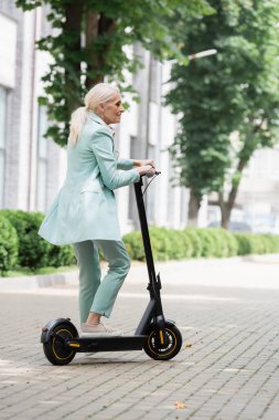 Mavi takım elbiseli üst düzey bir iş kadını şehrin caddesinde elinde elektrikli scooterla dikiliyor.
