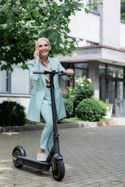 Elektrikli scooter 'ı olan yaşlı ve mutlu bir iş kadını dışarıda akıllı telefondan konuşuyor.