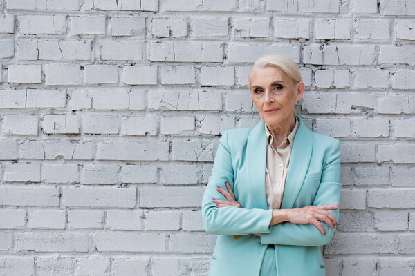 senior confident businesswoman standing with crossed arms near white brick wall