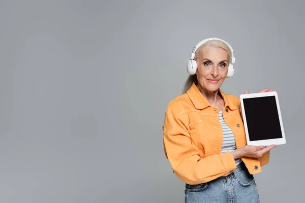 ヘッドフォンの流行のシニア女性は灰色に隔離された空白の画面でデジタルタブレットを示しています — ストック写真