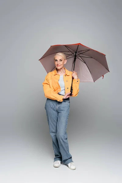 灰色の傘の下に立つジャケットとジーンズの幸せな高齢者の女性 — ストック写真