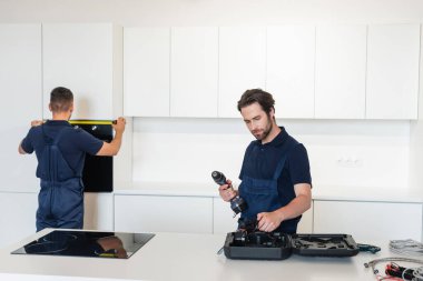 Meslektaşının yanında elektrikli matkap tutan işçiler mutfaktaki fırını ölçüyorlar.