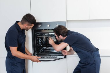 Tulumlu işçiler mutfakta elektrikli fırın tamir ediyorlar.