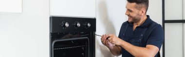 Modern mutfakta elektrikli fırını tamir eden mutlu tamirci, afiş.