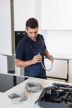 İşçi alet çantasının yanındaki metal hortuma ve mutfak masasındaki kablolara bakıyor.