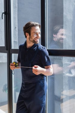 happy handyman with measuring tape looking away near large windows clipart