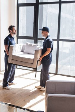 Üniformalı işçiler oturma odasında pencere kenarında koltuk taşıyorlar.