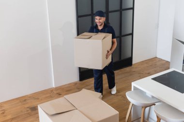 smiling mover carrying cardboard box in apartment clipart