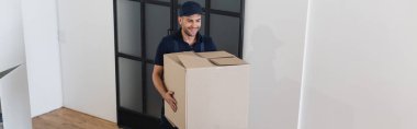 joyful mover in overalls carrying large carton box in apartment, banner clipart