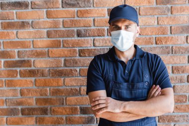 Tuğla duvarın yanında kolları çapraz duran tıbbi maskeli adam.