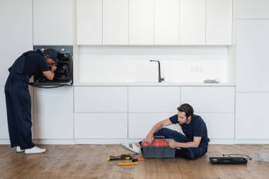 Mutfak zemininde alet çantasıyla oturan meslektaşının yanındaki elektrikli fırını tamir eden tamirci.