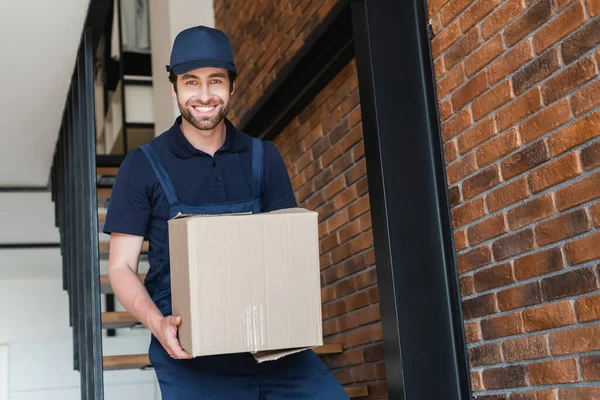 Karton paketle aşağıda yürürken kameraya gülümseyen teslimatçı.