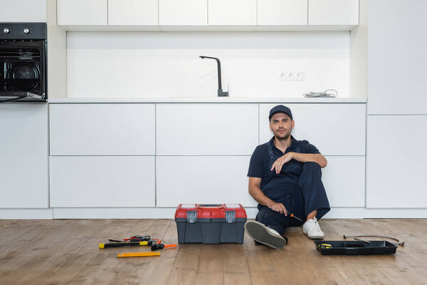 handyman in overalls sitting on floor in kitchen near tools and toolbox