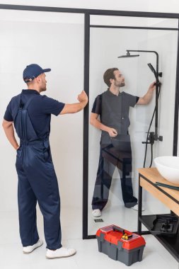 worker standing with hand on hip near plumber checking shower in bathroom clipart
