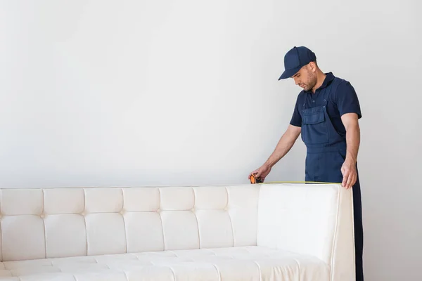 Trabalhador Boné Macacão Medindo Sofá Moderno Perto Parede Branca — Fotografia de Stock