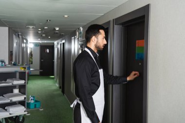 side view of bearded housekeeper knocking door in hotel room  clipart