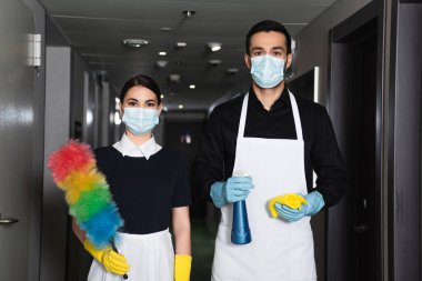 Tıbbi maskeli temizlikçiler ve lastik eldivenler ellerinde bez ve toz fırçasıyla sprey şişesi tutuyorlar.