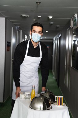 Tıbbi maskeli garson ve önlüklü servis tepsisi saat ve içeceklerle. 