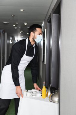 Tıbbi maskeli garson ve önlüklü servis tepsisi ve oteldeki içkiler. 