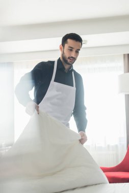 Sakallı hizmetçi otel odasında beyaz yatak değiştirip gülümsüyor.