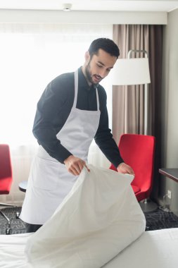 Gülümseyen ve sakallı hizmetçi otel odasında beyaz çarşaf değiştiriyor.