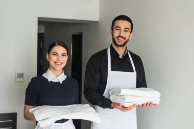 Gülümseyen temizlikçiler otel odasında beyaz çarşaf ve havlu tutuyorlar.