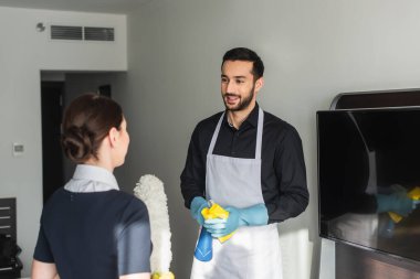 Toz fırçalı, bulanık oda hizmetçisine bakan lastik eldivenli neşeli hizmetçi.