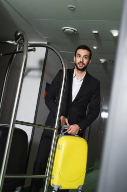 Gülümseyen komi otel koridorunda metal çan arabasının yanında valiz taşıyor.