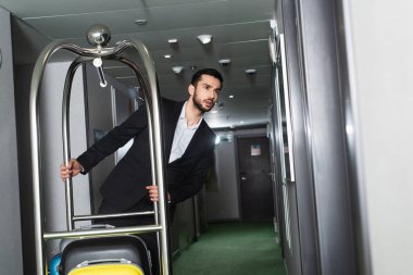 Sakallı belboy otelin koridorunda bavullu metal çan arabası itiyor.