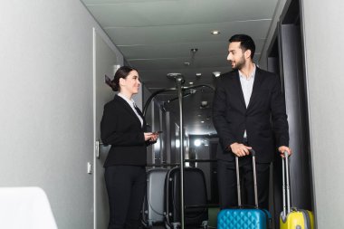 Sırıtkan resepsiyonist elinde akıllı telefon ve valizli komiye bakıyor. 