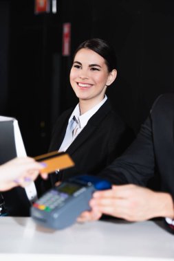 happy receptionist looking at tourist paying with credit card near colleague  clipart