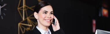 Otel lobisinde gülümseyen ve akıllı telefondan konuşan mutlu resepsiyonist.