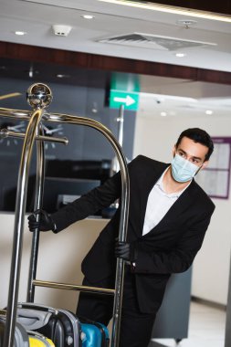 Sağlık maskeli komi otel lobisinde bagaj arabasını itiyor. 