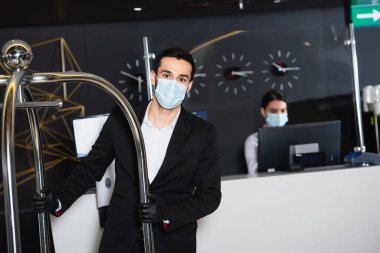 Sağlık maskeli komi, otel lobisindeki bulanık yöneticinin yanına bagaj arabasını itiyor. 