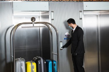 Otel asansörünün yanında duran sağlık maskeli komi. 