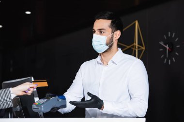 Tıbbi maske ve lateks eldivenli resepsiyonist kredi kartıyla turistin yanında ödeme terminali tutuyor. 