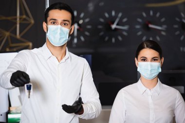Tıbbi maskeli resepsiyonist meslektaşının yanında bulanık oda anahtarını tutuyor. 