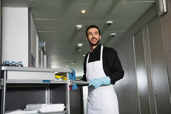 Fröhliche Und Bärtige Haushälterin Gummihandschuhen Die Neben Einkaufswagen Hotelflur Steht — Stockfoto