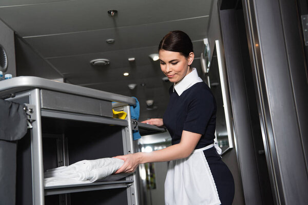 brunette maid in uniform taking clean bed sheets from housekeeping cart